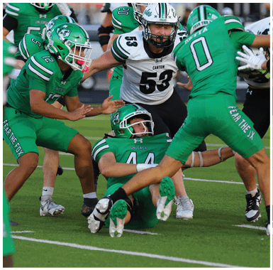 Four people in football uniforms are engaged in a play on a field, with one person in green lying on the ground while others in green and white surround them. Descripci n generada con IA