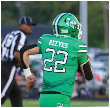 A person wearing a green football uniform with the name Reeves and number 22 is running on a football field with a referee and another player nearby. Descripci n generada con IA