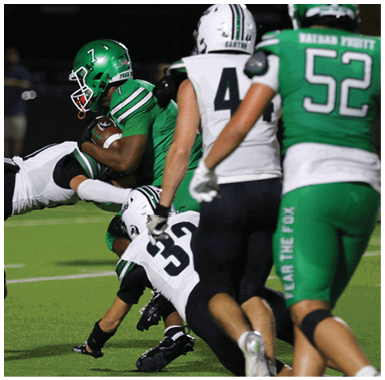 A person in a green football uniform is holding the ball while being tackled by two people in white and black uniforms on a football field. Descripci n generada con IA
