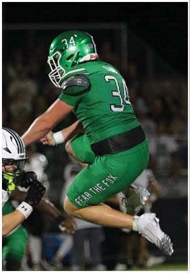 A person in a green football uniform with the number 34 is jumping over another person in a white football uniform during a nighttime game. Descripci n generada con IA
