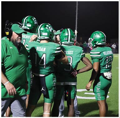 Three people in green football uniforms and helmets stand on a field at night with one person facing away from the camera and the others partially turned. Descripci n generada con IA