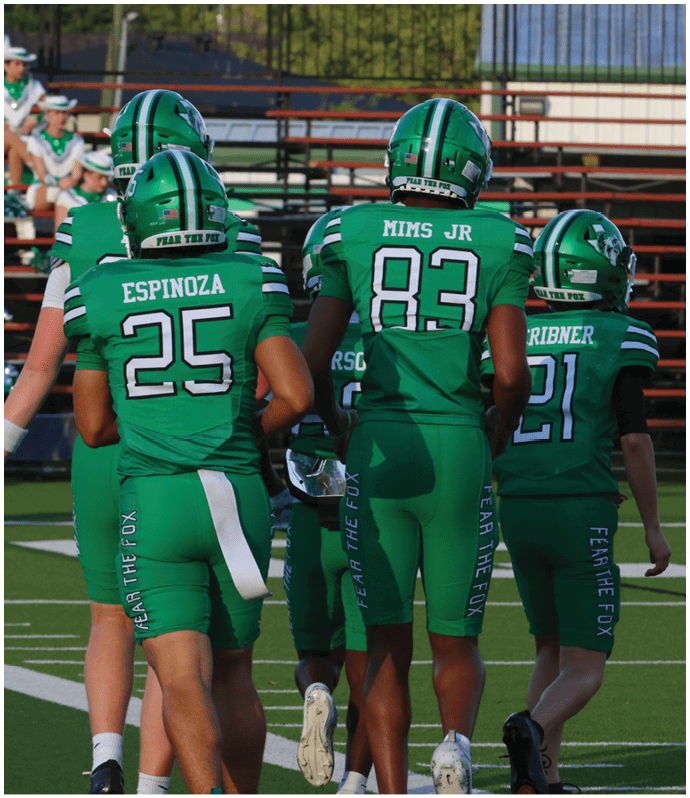 Four people in green football uniforms with helmets stand on a field with bleachers and cheerleaders in the background. Descripci n generada con IA