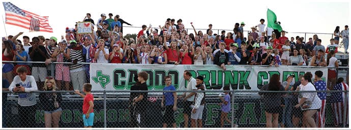 A crowd of people stands behind a fence in front of a large banner that reads “Caddo Trunk or Treat" with some people wearing casual clothing and others holding up phones or waving. Descripci n generada con IA