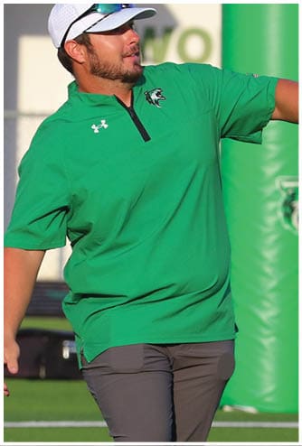 A person wearing a green shirt, gray pants, a white cap, and sunglasses is walking on a sports field with a tripod and equipment in the background. Descripci n generada con IA