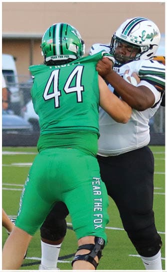 Two people in football uniforms are engaged in blocking on a football field with another player running in the background. Descripci n generada con IA
