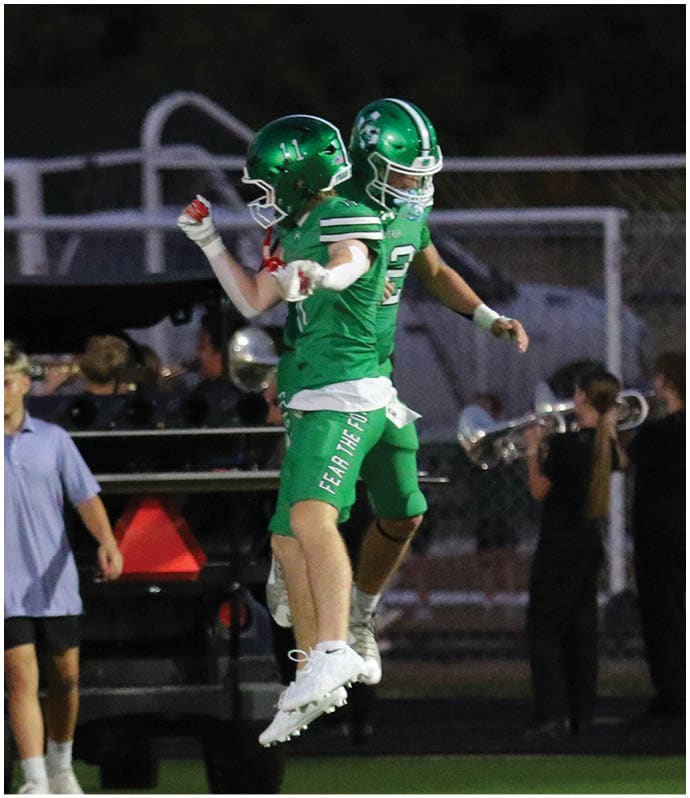 Two people in green football uniforms and helmets jump in celebration on a football field while others stand in the background near a golf cart and band members. Descripci n generada con IA