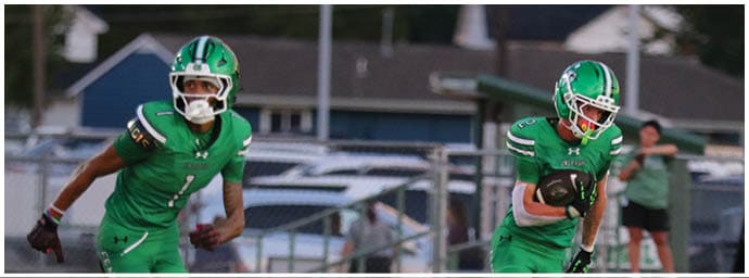 Two people in green football uniforms are running on a field during a game with a blurred background of spectators and equipment. Descripci n generada con IA