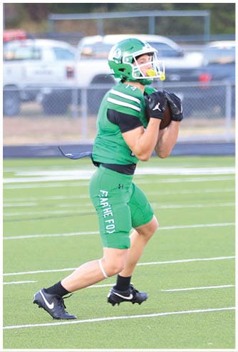 A person in green football gear and helmet is catching a football on a field with a blurred background of a fence, vehicles, and a sign. Descripci n generada con IA