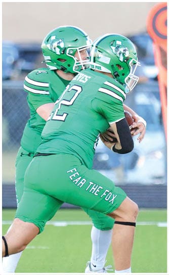 Two people in green football uniforms and helmets are engaged in a play on a football field with one person handing off the ball to the other. Descripci n generada con IA