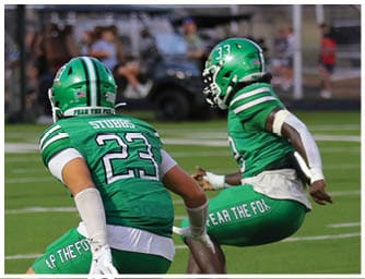 Two people in green football uniforms are crouched on a field while another person in a white and black uniform stands nearby during a game. Descripci n generada con IA