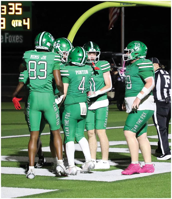 A group of football players in green uniforms are gathered on the field near the end zone while a referee stands in the foreground and the scoreboard shows the home team leading. Descripci n generada con IA