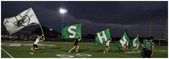 People running on a football field at dusk holding large flags with letters and a mascot logo. Descripci n generada con IA