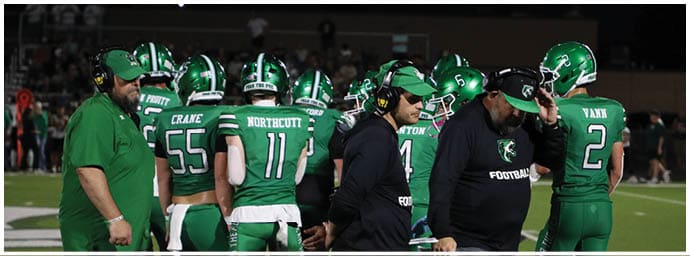 A group of people wearing green football uniforms and helmets are gathered on a field with two adults in black coaching attire standing among them. Descripci n generada con IA