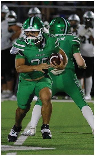 Two people in green football uniforms are on a field with one holding a football and the other preparing to receive it while players in white and black uniforms stand in the background. Descripci n generada con IA