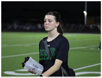 A person with long hair tied back wearing a black sports shirt and carrying a bag is holding a water bottle while standing on a sports field at night. Descripci n generada con IA