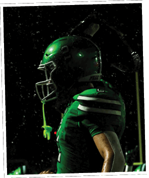 A person wearing green football pants with the words “FEAR THE FOX" printed on the side is preparing to throw a football on a turf field. Descripci n generada con IA