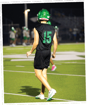 A person wearing a green helmet and black sports uniform with the number 15 is standing on a football field at night. Descripci n generada con IA