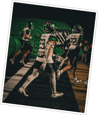 People wearing football uniforms and helmets are running onto a field from a large inflatable tunnel at night. Descripci n generada con IA