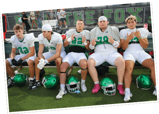 Three people in football uniforms sit on a bench on a field with helmets on the ground in front of them, making hand gestures and looking at the camera. Descripci n generada con IA