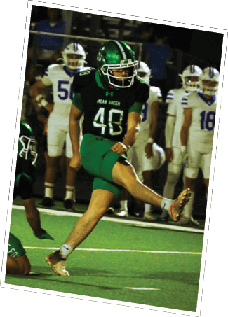 A person in a green and black football uniform is kicking a ball while another person in a similar uniform is crouched nearby, with people in white uniforms standing in the background. Descripci n generada con IA