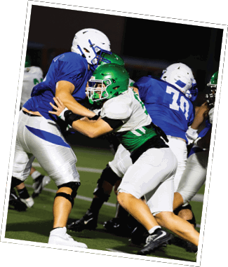 Two people in football uniforms are engaged in a blocking action on a field during a game at night. Descripci n generada con IA