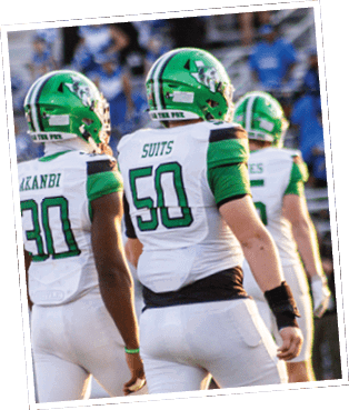 People wearing green and white football uniforms with helmets are standing on a field with blurred spectators in the background. Descripci n generada con IA