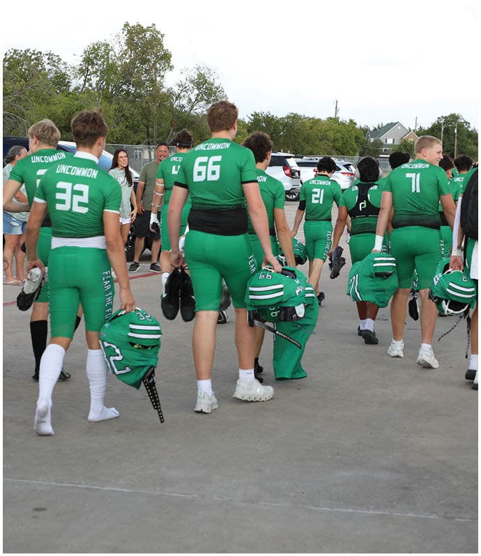 A group of people wearing green sports uniforms walk away on a paved surface carrying helmets and bags, while one person on the right side holds a phone to take a photo or video. Descripci n generada con IA