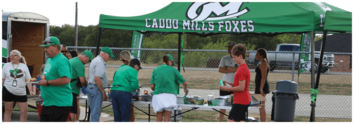 People wearing green shirts and hats are gathered under a green canopy labeled “CADDO MILLS FOXES" serving food and drinks to others near a chain-link fence. Descripci n generada con IA
