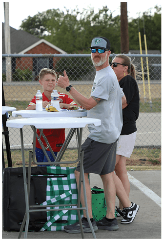 An adult wearing a cap and sunglasses gives a thumbs-up while sitting at a table with a child in a sports jersey and another adult standing nearby outdoors. Descripci n generada con IA