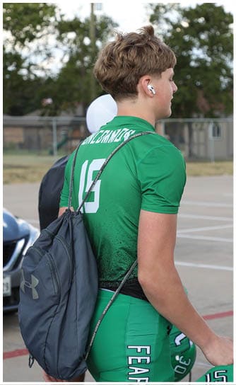A person wearing a green sports uniform with the number 15 and a drawstring backpack is standing outdoors with wireless earbuds in their ears. Descripci n generada con IA