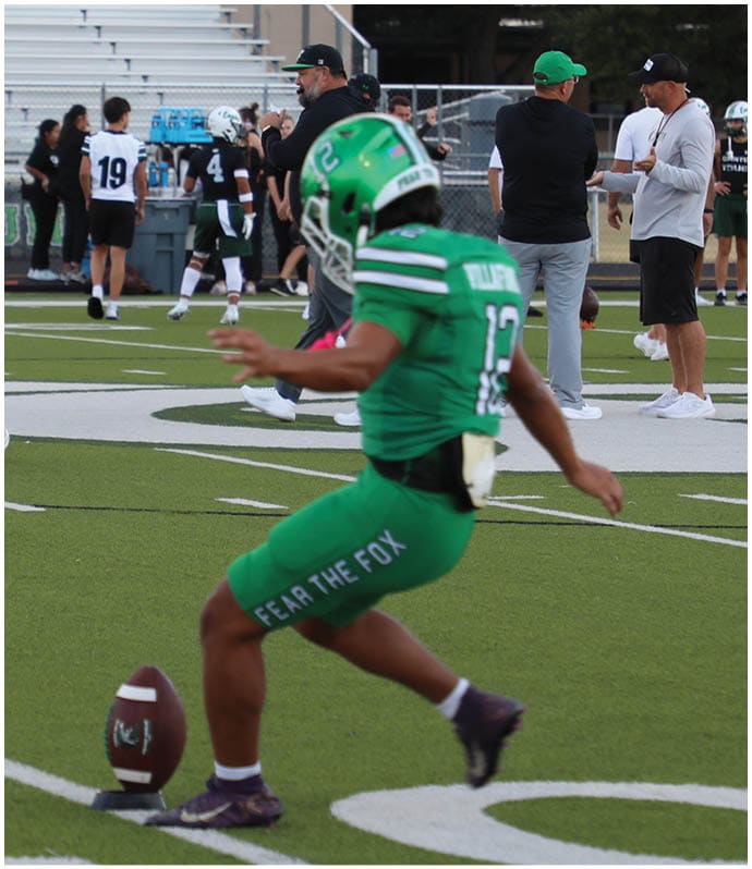 A person in green sports gear is kicking a football on a field while several people stand and walk in the background near bleachers. Descripci n generada con IA