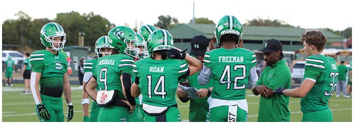 A group of people wearing green football uniforms and helmets are gathered on a football field, appearing to discuss strategy or receive instructions. Descripci n generada con IA