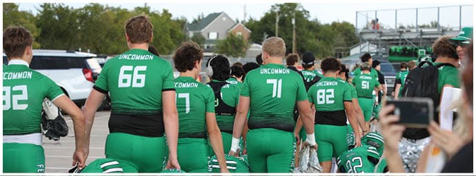 People wearing green football uniforms with the word “UNCOMMON" on the back are walking on a field with some holding helmets and others carrying equipment. Descripci n generada con IA