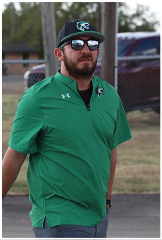 A person wearing a green sports shirt and a black cap with a logo stands outdoors near a chain-link fence and parked vehicles. Descripci n generada con IA