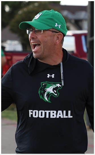 A person wearing a green cap and a black hoodie with a wolf logo and the word “FOOTBALL" is smiling and holding a water bottle outdoors. Descripci n generada con IA