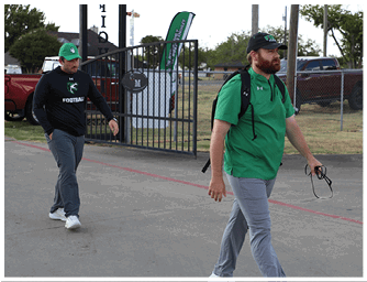 A person wearing a green shirt, gray pants, a black cap, and a backpack is walking on a paved area near a chain-link fence and a parked truck. Descripci n generada con IA