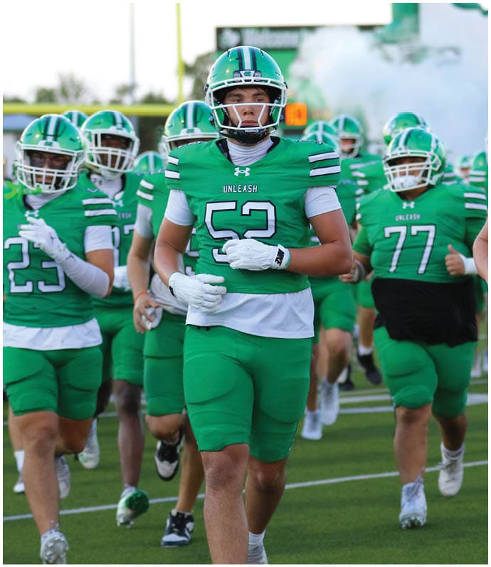 A group of people wearing green football uniforms with helmets are running onto a field with smoke in the background. Descripci n generada con IA