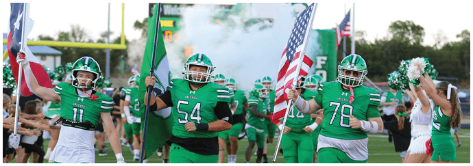 People in green football uniforms run on a field holding flags while cheerleaders in green and white outfits cheer on the sidelines. Descripci n generada con IA