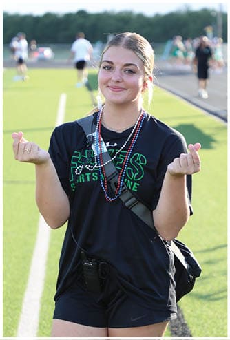 A person wearing a black shirt and beaded necklaces is making a finger heart gesture outdoors on a sunny day. Descripci n generada con IA