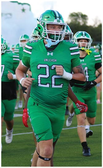 People in green football uniforms and helmets are running onto a field with smoke in the background. Descripci n generada con IA