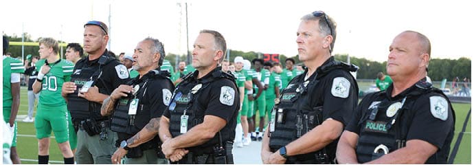 Several people in police uniforms stand in a line on a sports field with athletes in green uniforms visible in the background. Descripci n generada con IA