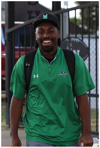 A person wearing a green shirt and carrying a backpack is holding a plastic water bottle near a black metal fence with a chain-link background. Descripci n generada con IA