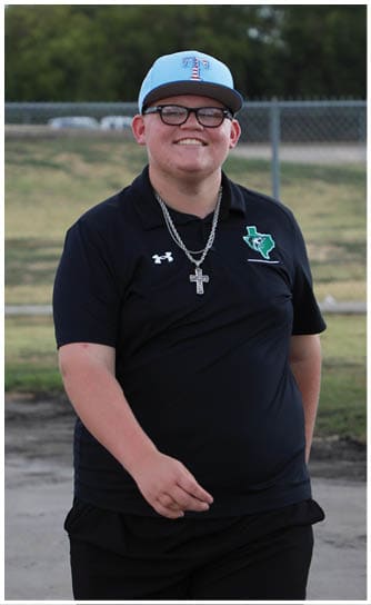 A person wearing glasses, a light blue baseball cap, a black collared shirt with a Texas emblem, and a silver chain necklace is smiling outdoors with trees and a fence in the background. Descripci n generada con IA