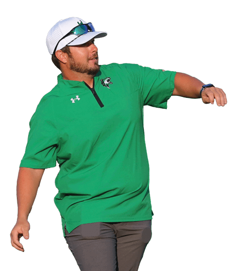A person wearing a green shirt, dark pants, white shoes, and a white cap is standing on a sports field with one foot slightly raised and arms extended. Descripci n generada con IA
