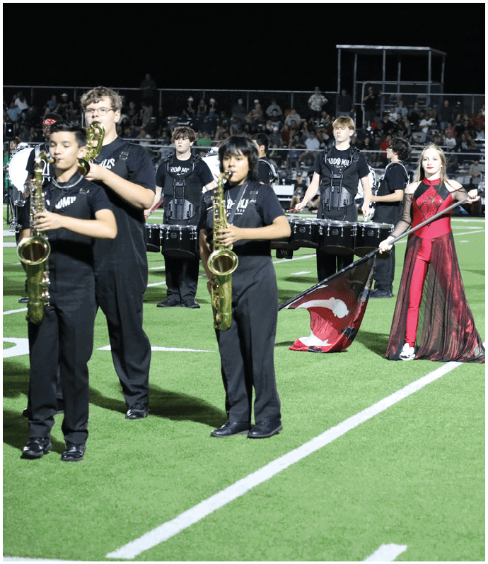 People in red and black costumes are performing a color guard routine with flags on a football field at night while a crowd watches from the bleachers. Descripci n generada con IA