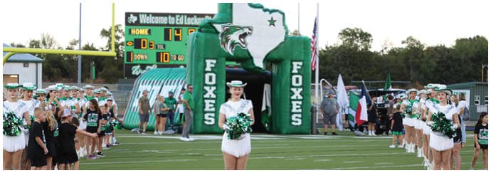 A person in a white uniform holding pom-poms stands on a football field with a group of people and cheerleaders lined up on the sidelines near a green inflatable tunnel labeled “FOXES." Descripci n generada con IA