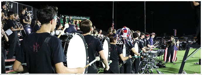 A group of people wearing black shirts are playing percussion instruments in a nighttime outdoor setting, with one person dressed in a costume resembling the American flag. Descripci n generada con IA