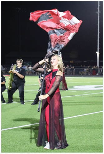 A person wearing a dark outfit is holding a large red and black flag on a sports field at night with a crowd of people in the background. Descripci n generada con IA