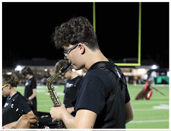 Person with curly hair and glasses playing a saxophone on a sports field at night. Descripci n generada con IA