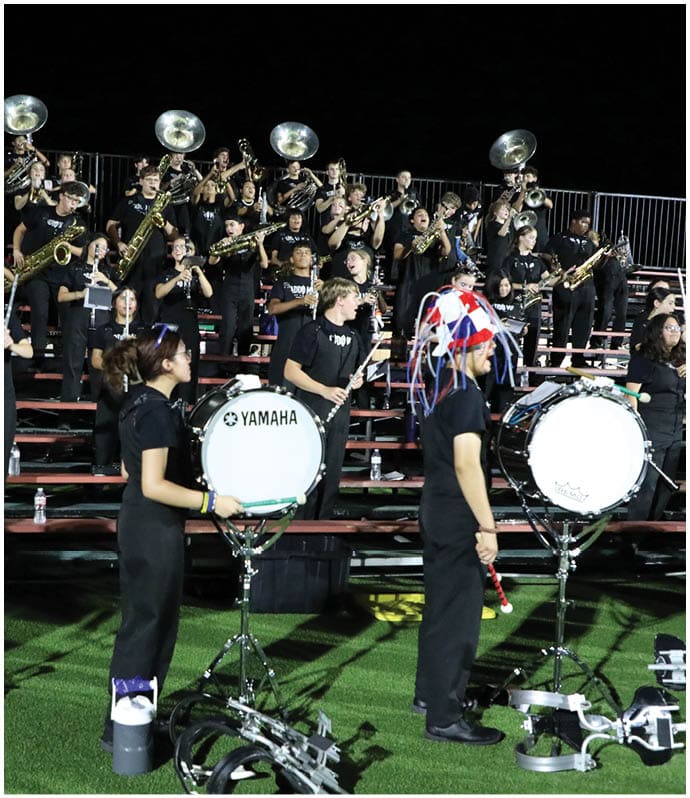 People in matching black uniforms are playing various musical instruments in a band setting on bleachers at night. Descripci n generada con IA